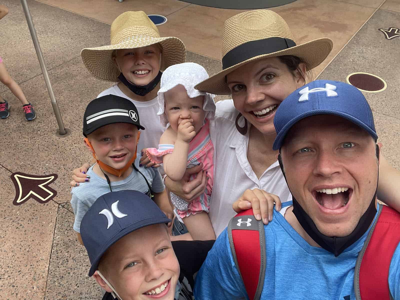 Selfie of Lisa P.’s family at Animal Kingdom — her husband and three kids gathered closely with a baby in the middle, all wearing hats and smiling excitedly.