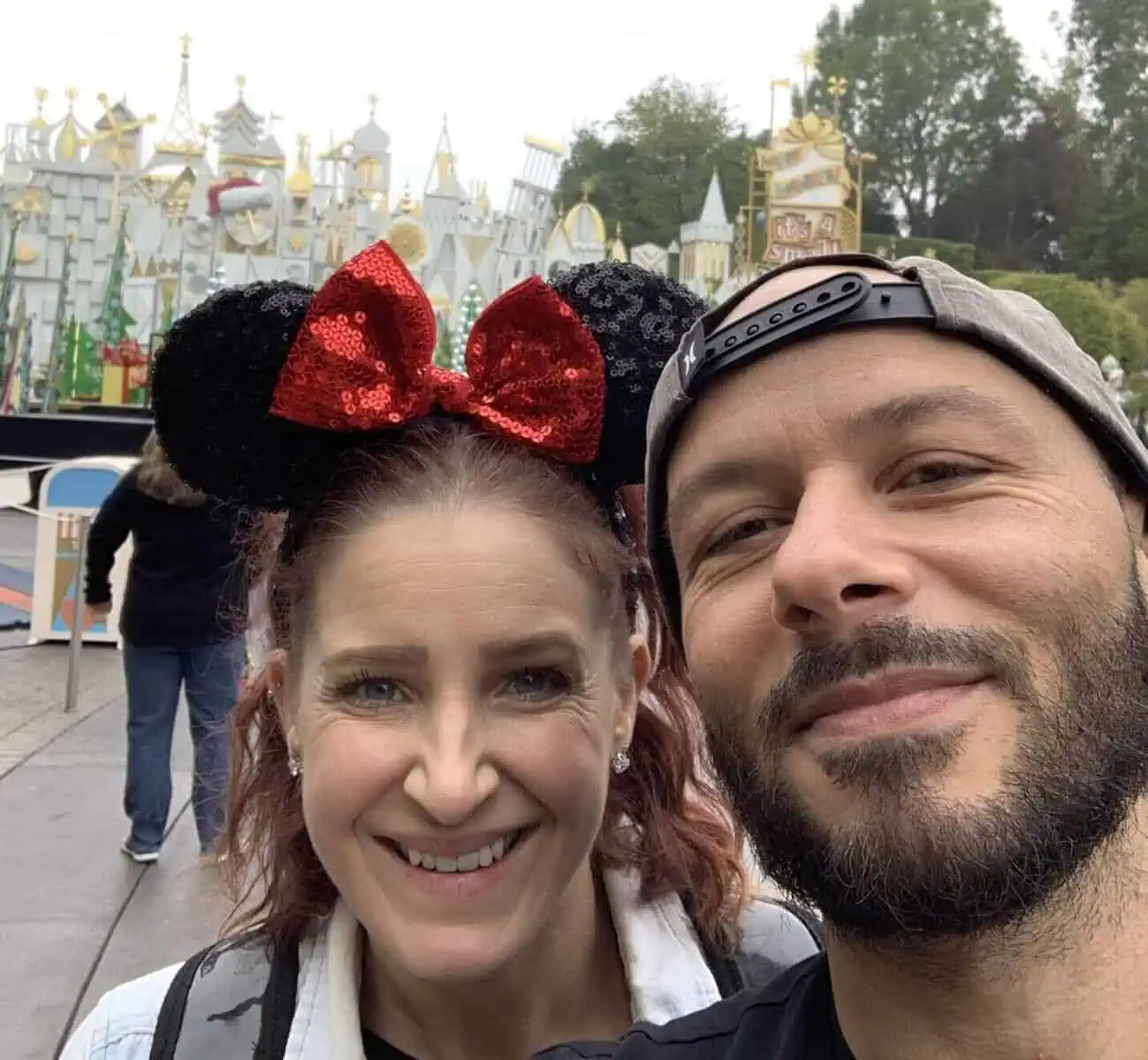 Selfie of Maggie B. and her husband on their Disneyland honeymoon, with Maggie wearing Minnie ears and the façade of It’s a Small World in the background.