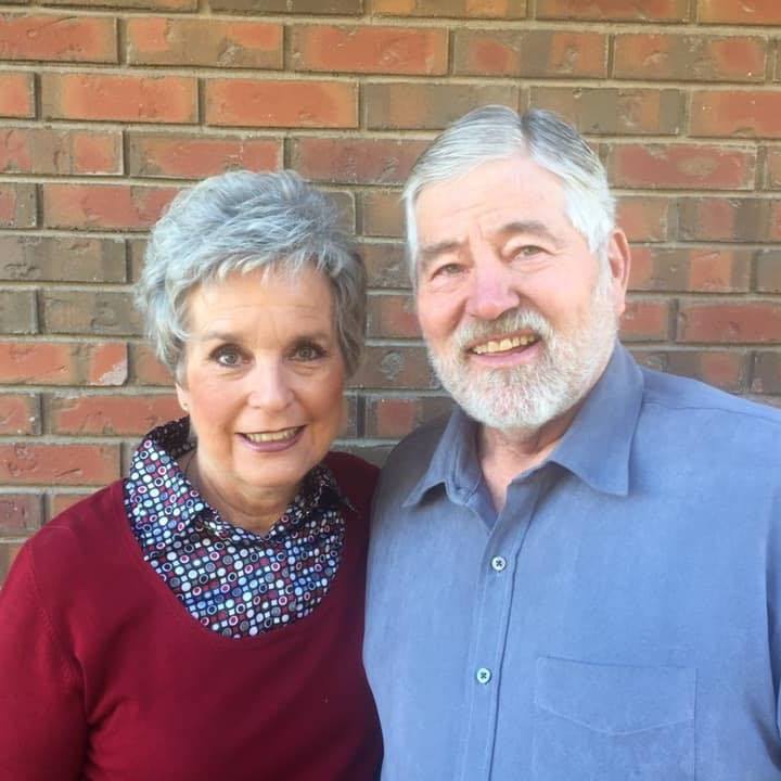 Photo of Miriam A. and her husband standing by a brick wall, smiling — they appear to be proud and happy grandparents.