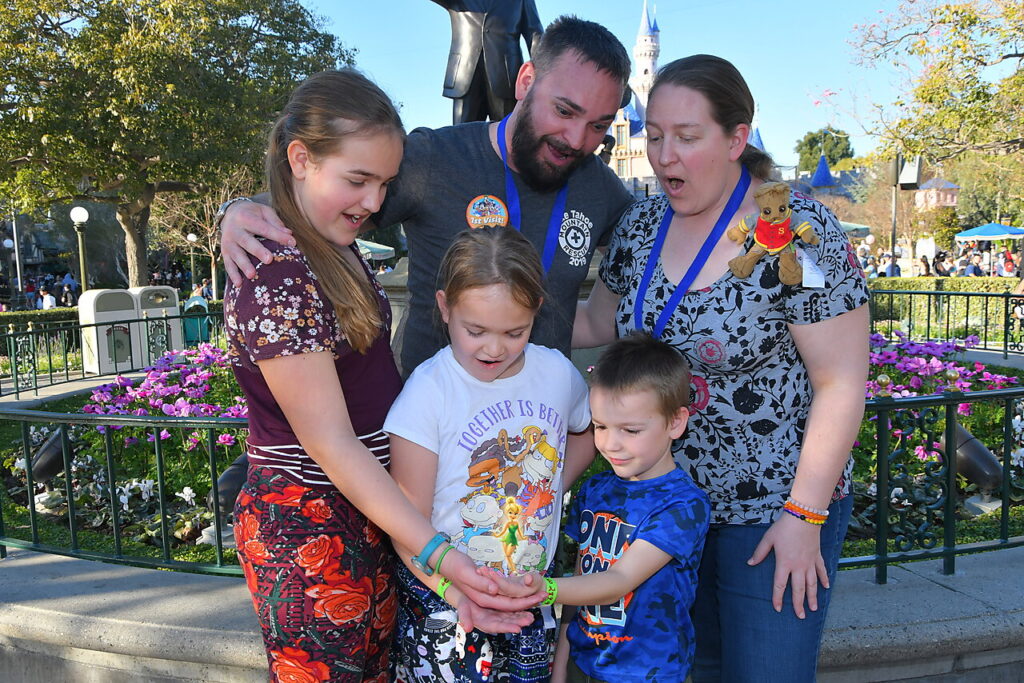 Photo of Richard M.’s family at Disneyland in front of the castle, standing in a half circle with surprised smiles as their three children hold out their hands with Tinker Bell standing in them.
